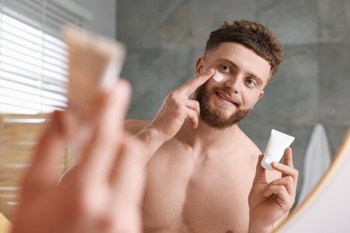 Handsome man applying moisturizing cream onto his face near mirror in bathroom