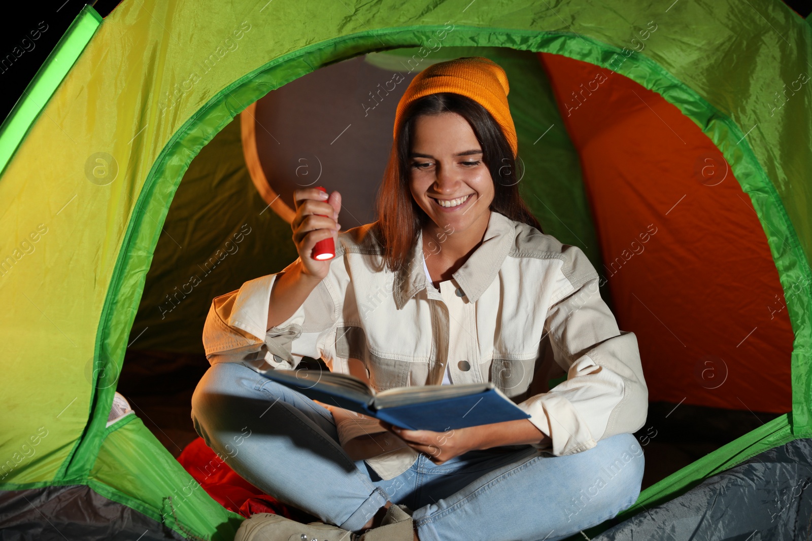 Photo of Young woman with flashlight reading book in tent at night