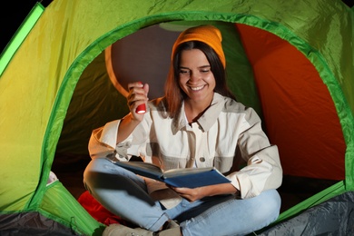 Young woman with flashlight reading book in tent at night