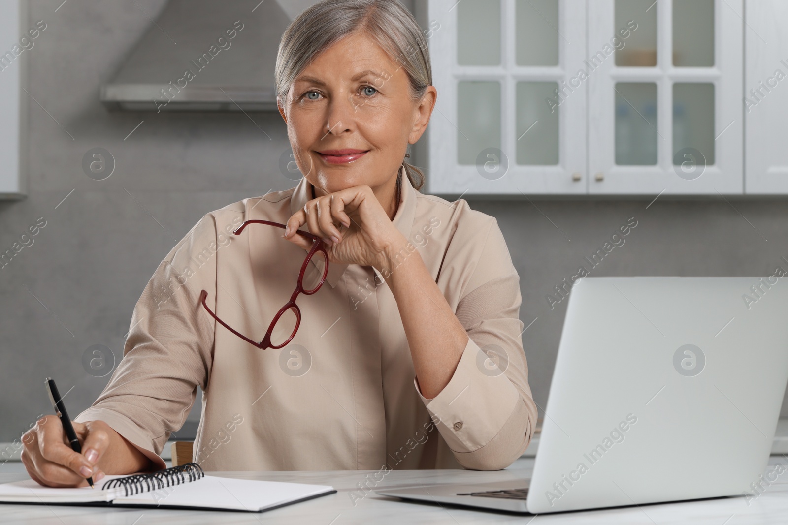 Photo of Beautiful senior woman taking notes near laptop at white marble table in kitchen