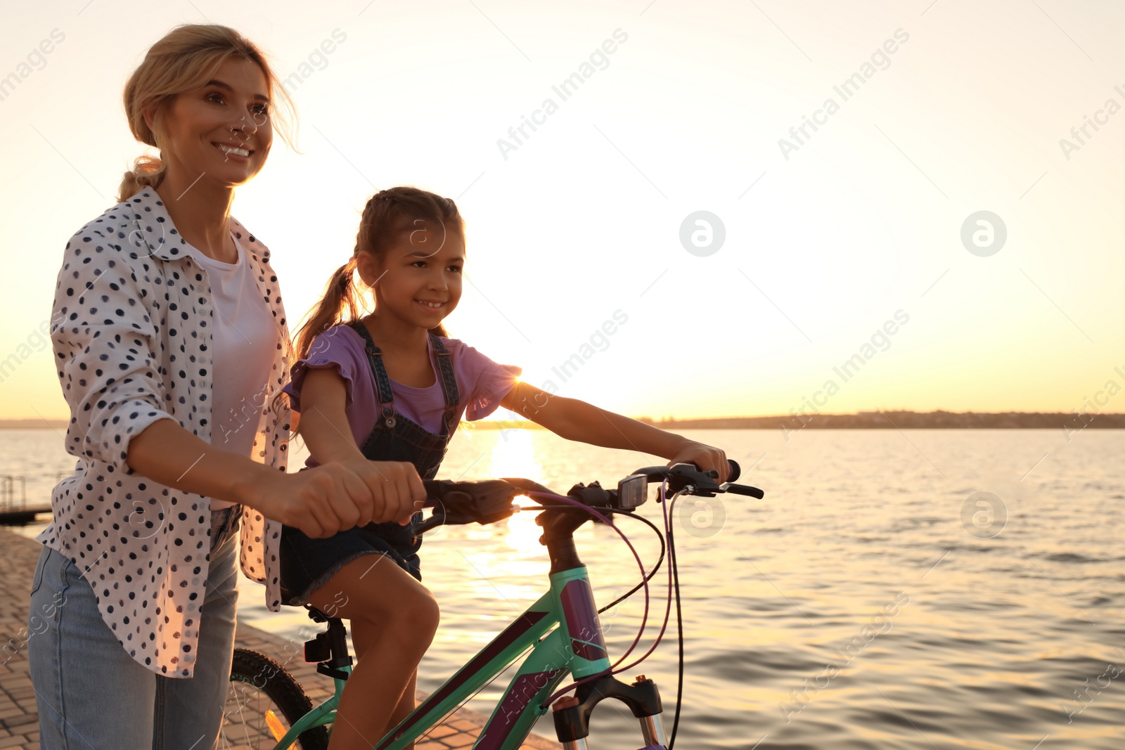 Photo of Happy mother teaching her daughter to ride bicycle near river at sunset