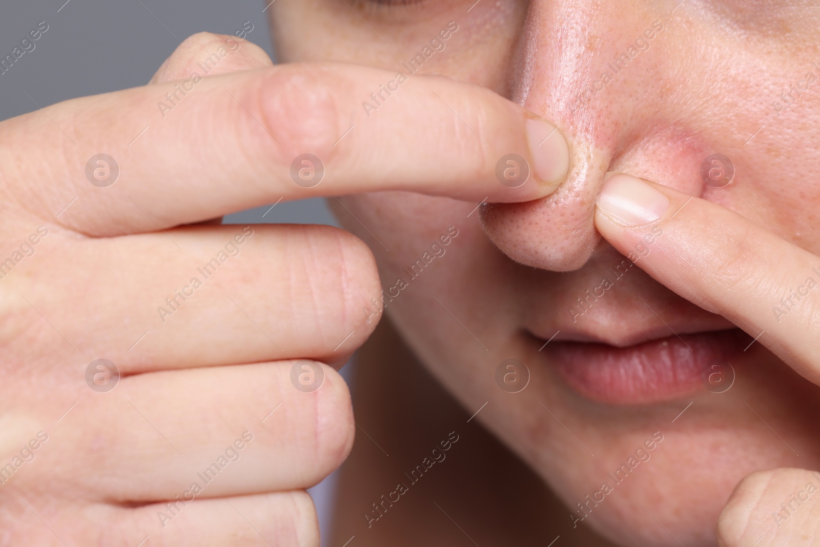 Photo of Woman popping pimple on her nose against grey background, closeup