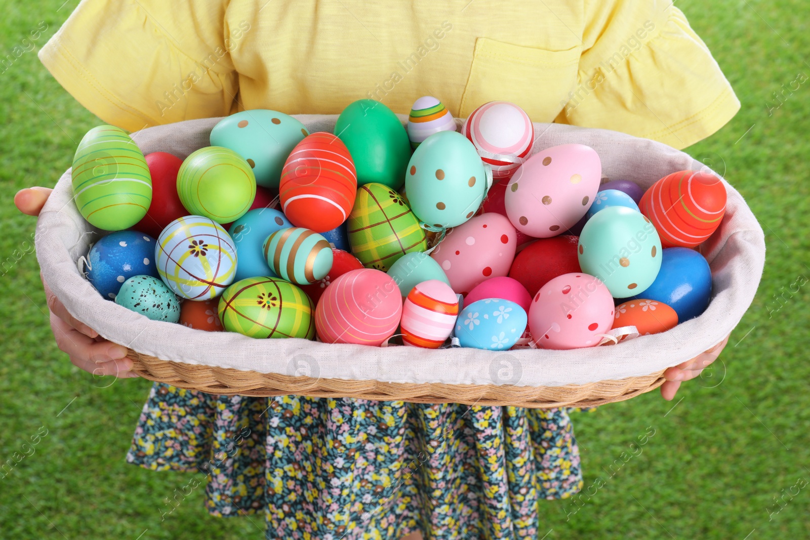 Photo of Little girl holding wicker basket full of Easter eggs outdoors, closeup