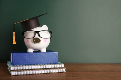 Photo of Piggy bank with graduation hat and books on table near chalkboard. Space for text