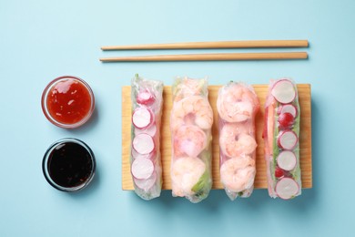 Different delicious spring rolls, chopsticks and sauces on light blue background, flat lay