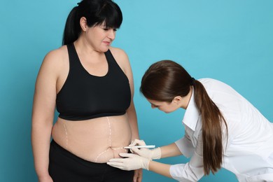Photo of Doctor drawing marks on obese woman's body against light blue background. Weight loss surgery