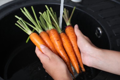 Photo of Woman washing ripe carrots with running water in sink, closeup