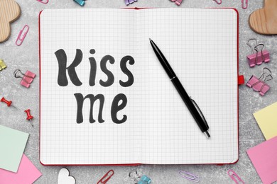 Photo of Phrase Kiss me written in notebook and stationery on light grey stone table, flat lay