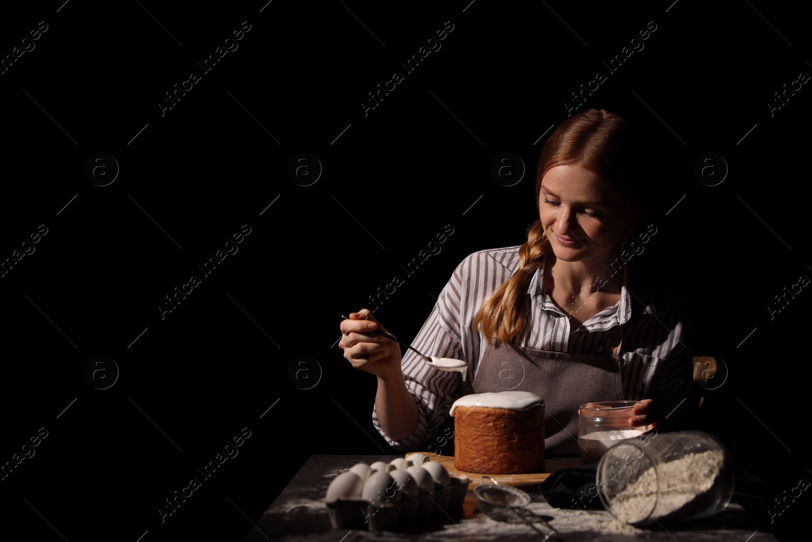 Photo of Young woman decorating traditional Easter cake with glaze at table against black background. Space for text