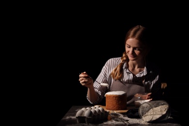 Photo of Young woman decorating traditional Easter cake with glaze at table against black background. Space for text