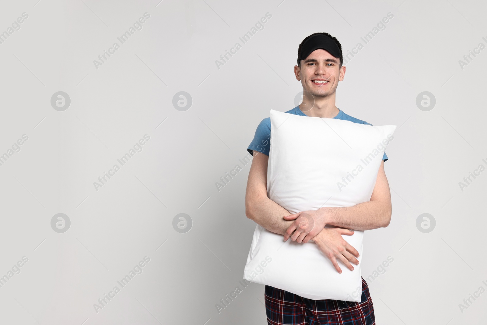 Photo of Happy man in pyjama and sleep mask holding pillow on light grey background, space for text