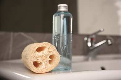 Natural loofah sponge and bottle with shower gel on washbasin in bathroom, closeup