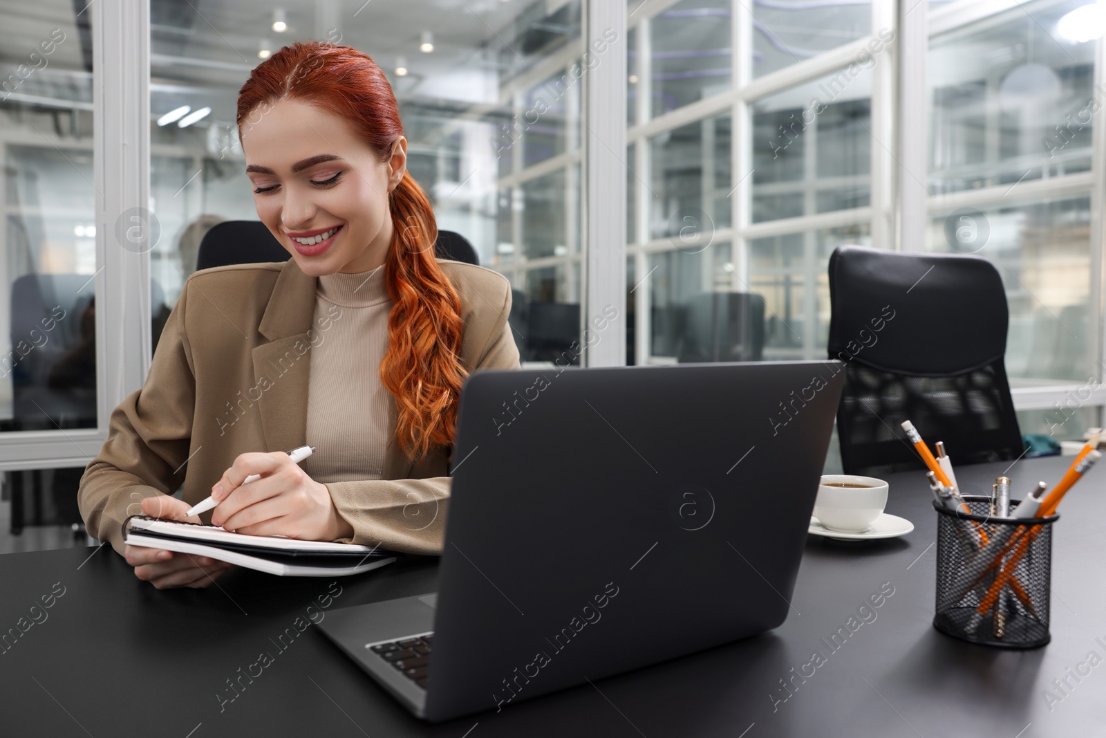 Photo of Happy woman taking notes while working with laptop at black desk in office