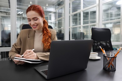 Photo of Happy woman taking notes while working with laptop at black desk in office