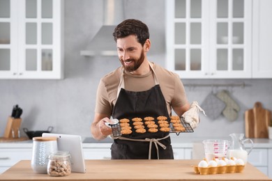 Photo of Man holding grid with freshly baked cookies while watching online cooking course via tablet in kitchen