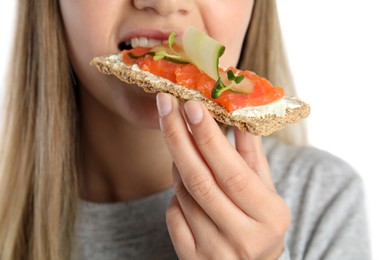 Photo of Woman eating fresh rye crispbread with cream cheese, salmon and cucumber on white background, closeup