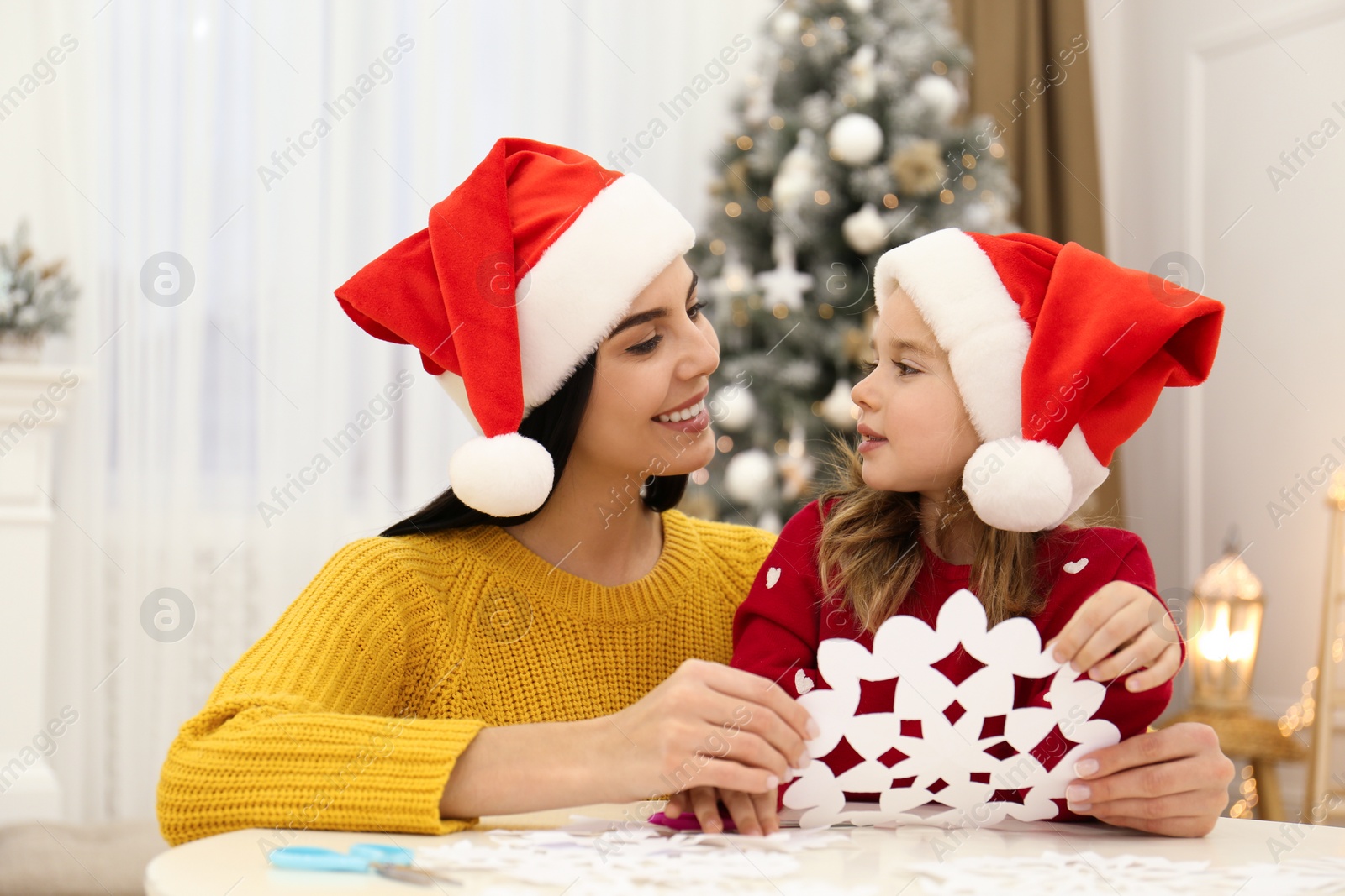 Photo of Mother and daughter in Santa hats with paper snowflake near Christmas tree at home