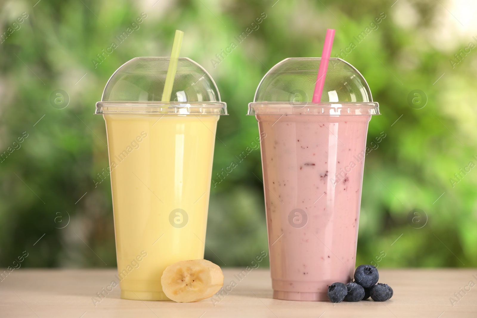 Photo of Plastic cups with different tasty smoothies and fresh fruits on wooden table outdoors