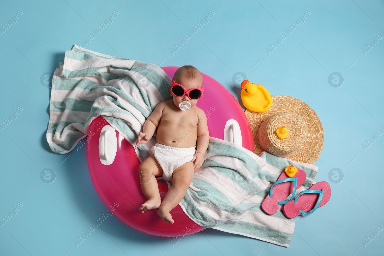 Photo of Cute little baby in sunglasses with inflatable ring and beach accessories on light blue background, top view