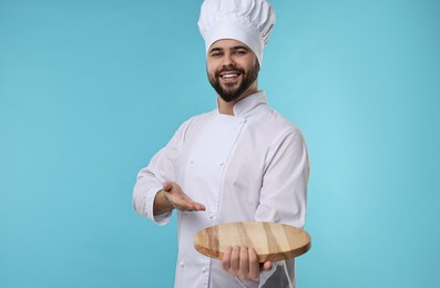 Photo of Happy young chef in uniform holding wooden board on light blue background