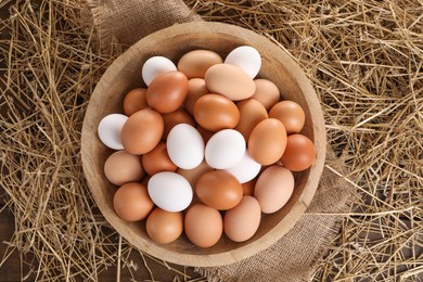 Fresh chicken eggs in bowl and dried straw on wooden table, top view