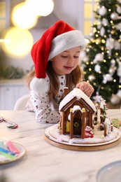 Photo of Cute little girl decorating gingerbread house at table indoors