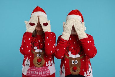 Photo of Senior couple in Christmas sweaters and Santa hats covering faces with hands in knitted mittens on light blue background