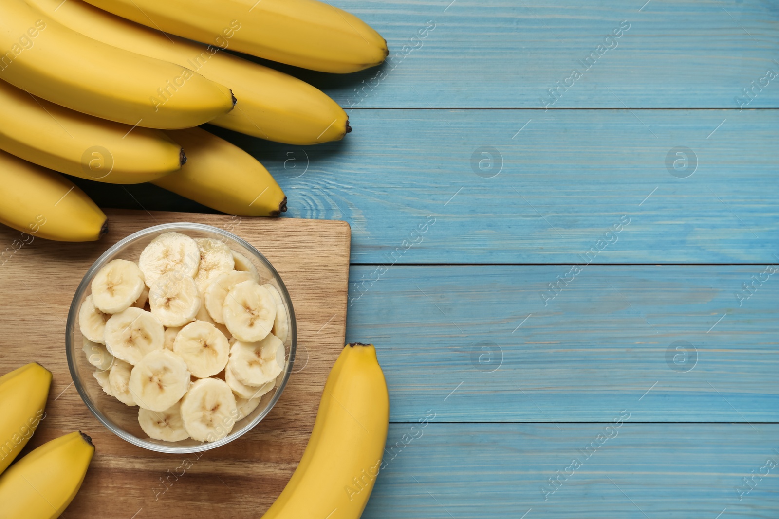 Photo of Bowl with cut bananas near whole fruits on light blue wooden table, flat lay. Space for text