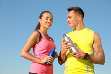 Young sporty couple with water bottles against blue sky on sunny day