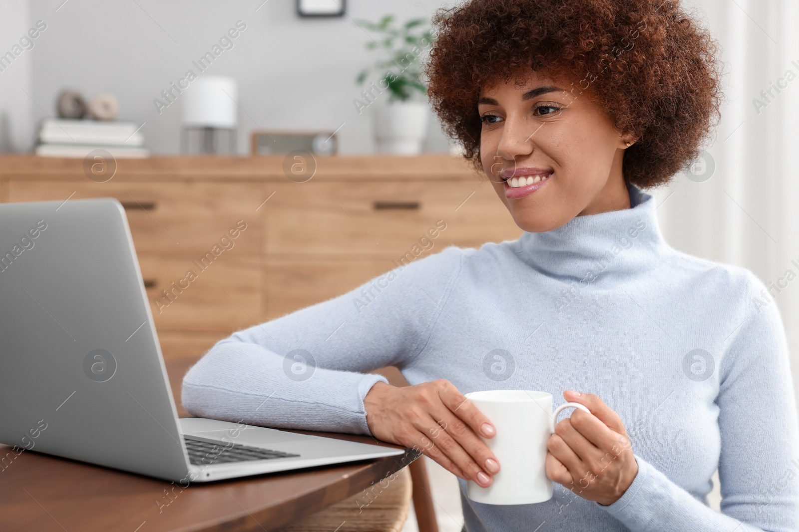 Photo of Beautiful young woman using laptop and drinking coffee at wooden table in room