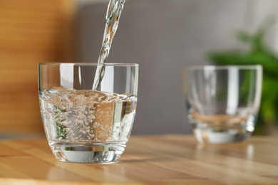 Photo of Pouring water into glass on wooden table indoors, space for text. Refreshing drink
