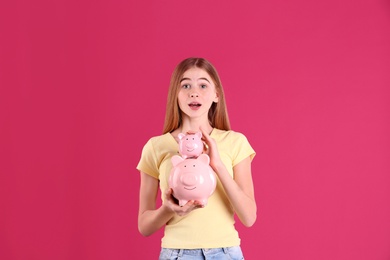 Emotional teen girl with piggy bank on color background