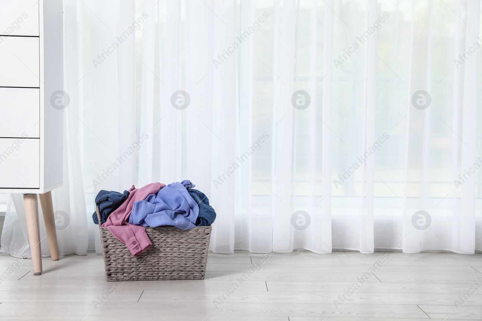 Photo of Wicker laundry basket with dirty clothes near window in room. Space for text