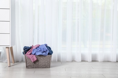 Photo of Wicker laundry basket with dirty clothes near window in room. Space for text