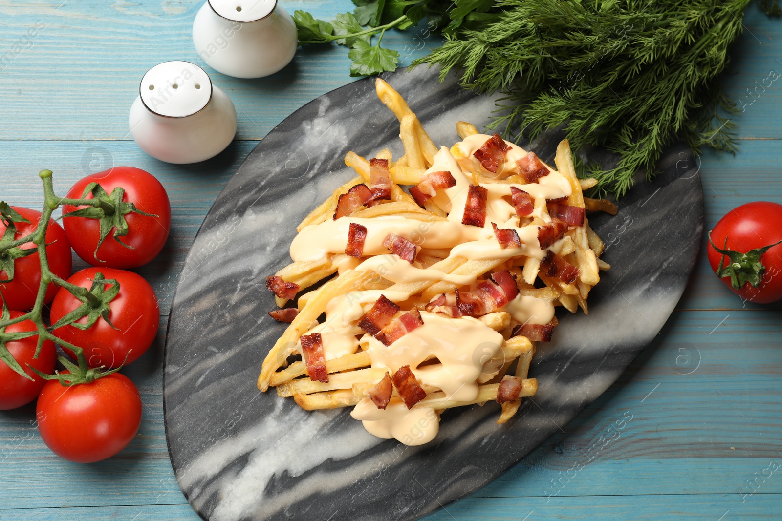 Photo of Delicious French fries with bacon, cheese sauce, tomatoes and herbs on light blue wooden table, flat lay