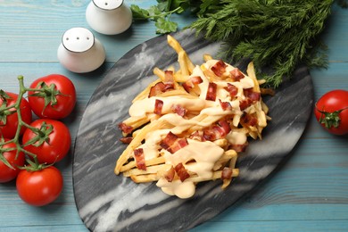 Delicious French fries with bacon, cheese sauce, tomatoes and herbs on light blue wooden table, flat lay