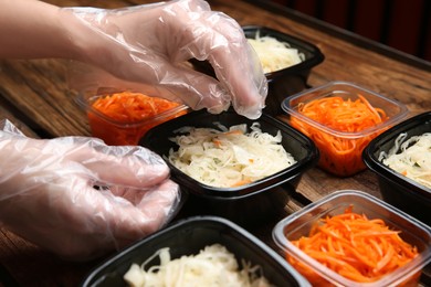 Photo of Waiter in gloves putting salads into containers at wooden table, closeup. Food delivery service