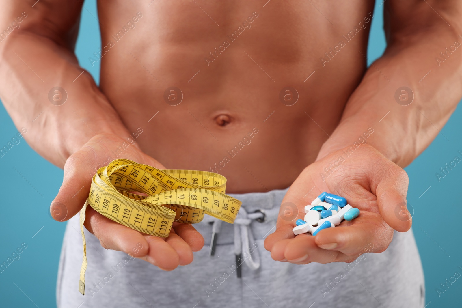 Photo of Athletic man with measuring tape and pills on light blue background, closeup. Weight loss