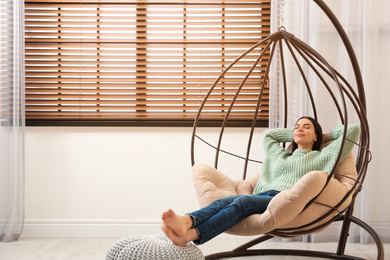 Young woman relaxing in hanging chair near window at home. Space for text