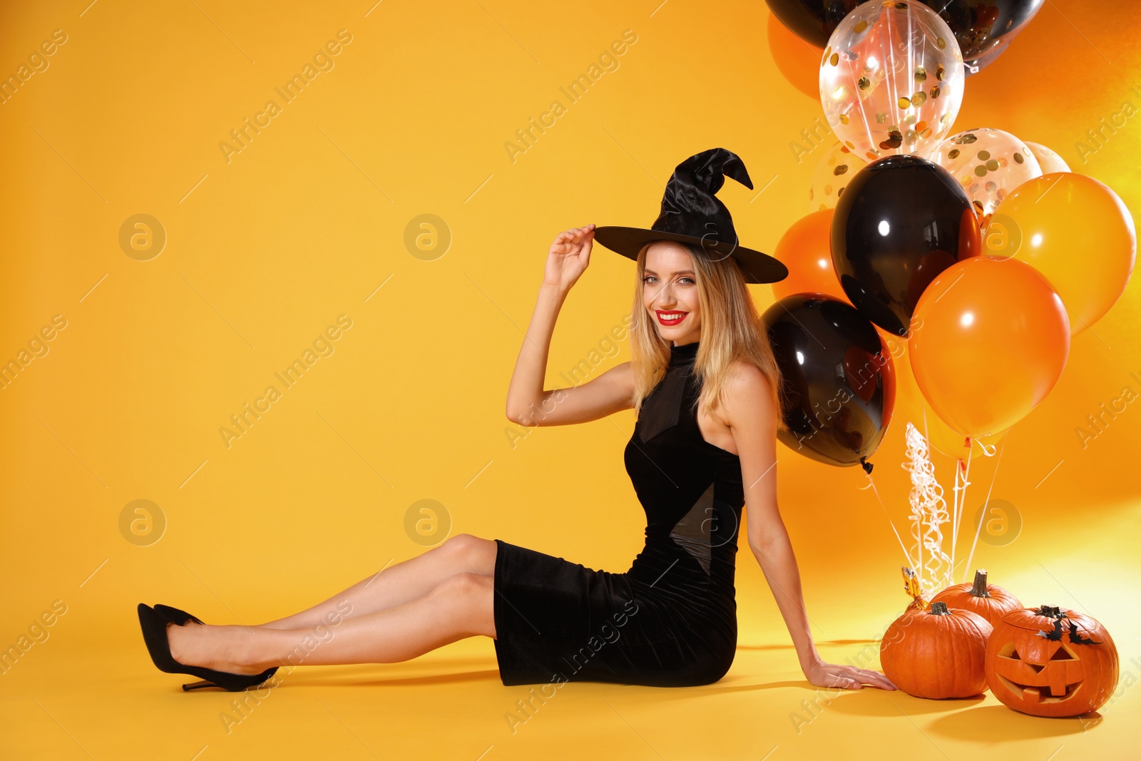 Photo of Beautiful woman in witch costume with balloons and pumpkins on yellow background, space for text. Halloween party