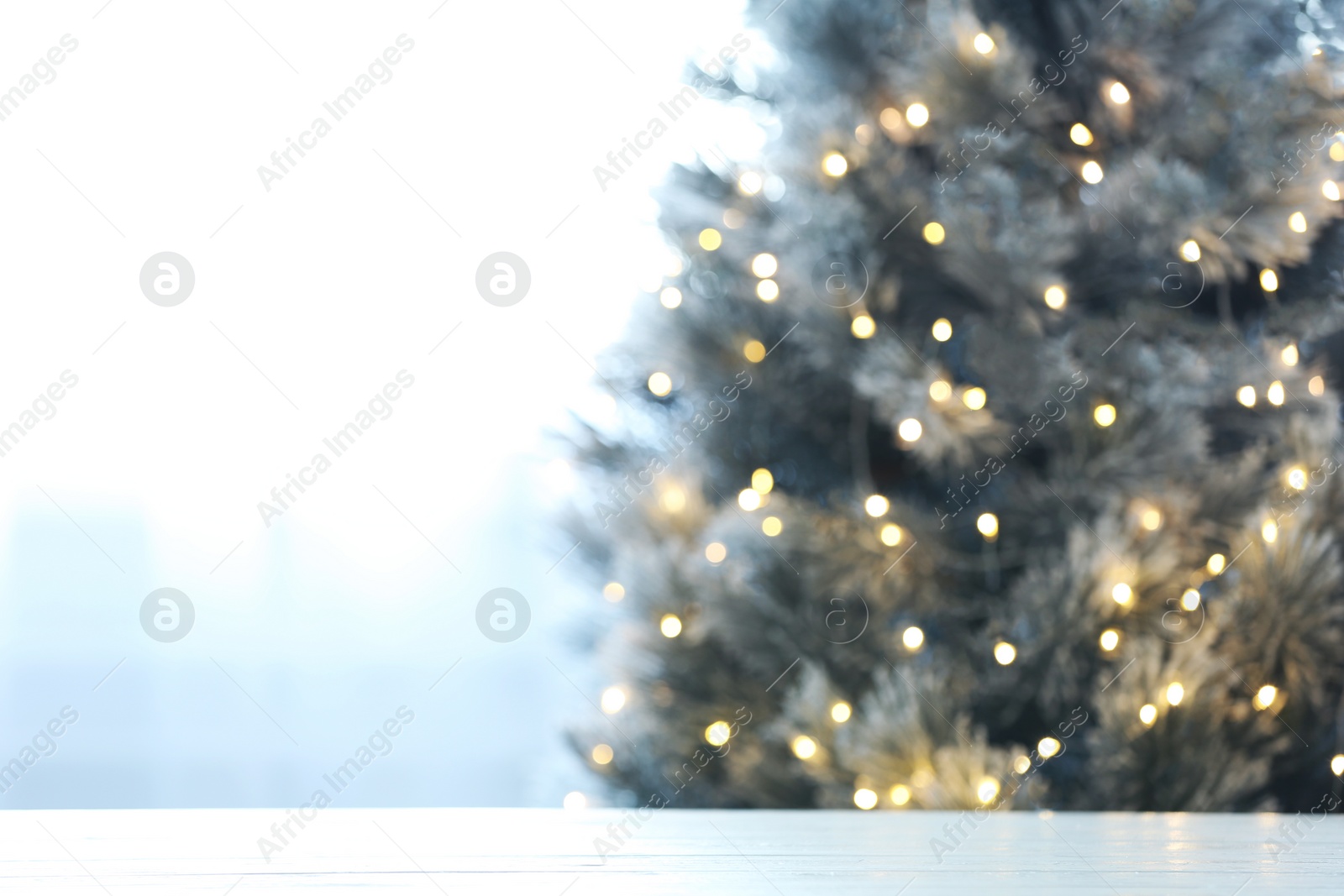 Photo of Blurred view of beautiful Christmas tree with yellow lights near window indoors, focus on wooden table. Space for text