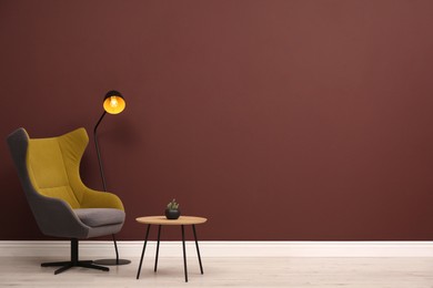 Photo of Stylish armchair, table and lamp near brown wall, space for text
