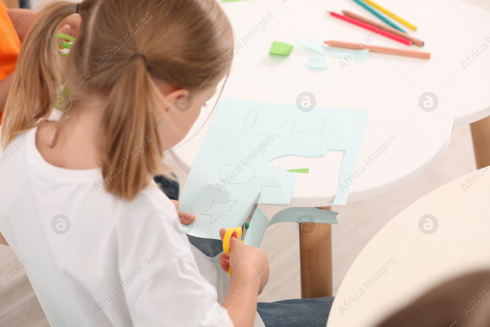 Photo of Little girl cutting color paper with scissors at desk, closeup. Kindergarten activities