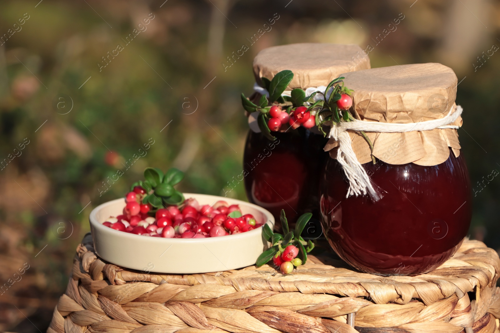 Photo of Jars of delicious lingonberry jam and red berries on wicker basket outdoors