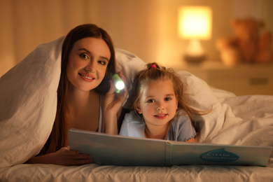 Photo of Mother and daughter with flashlight reading book under blanket at home