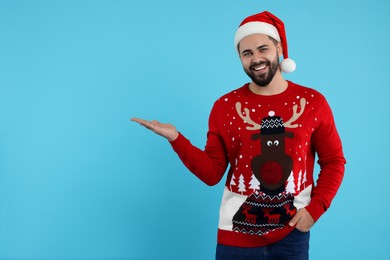 Happy young man in Christmas sweater and Santa hat showing something on light blue background. Space for text