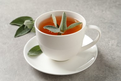 Photo of Cup of aromatic sage tea and fresh leaves on grey table