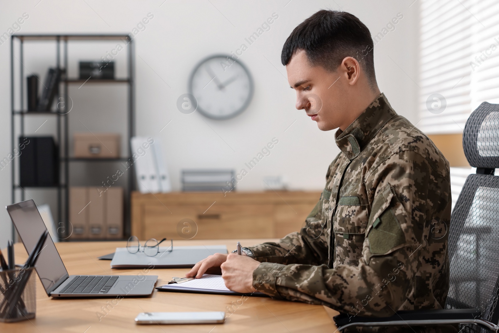 Photo of Military service. Young soldier working at wooden table in office