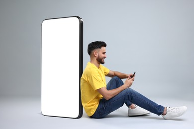 Man with mobile phone sitting near huge device with empty screen on grey background. Mockup for design