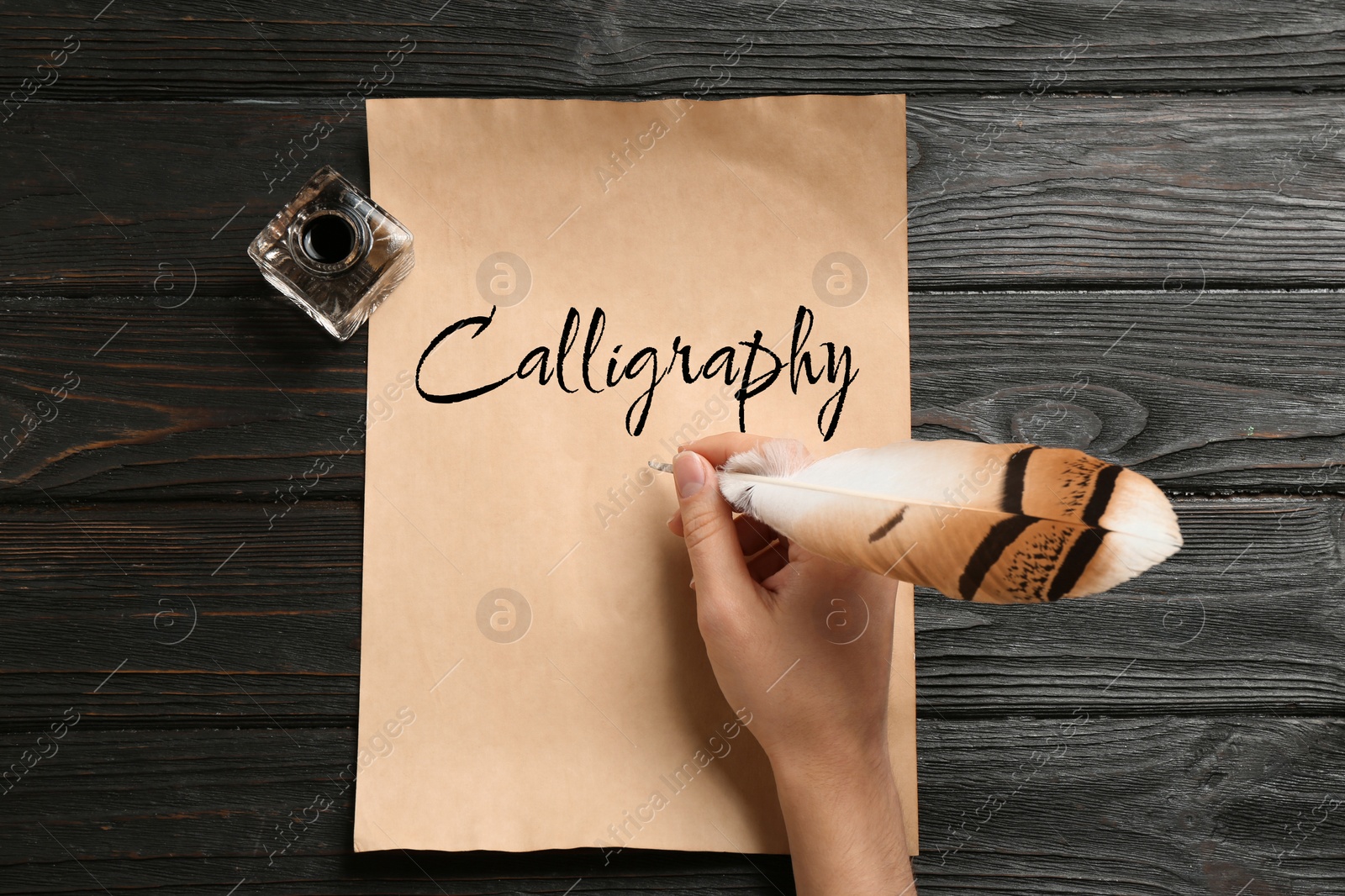 Image of Woman writing word Calligraphy with feather and ink on parchment at wooden table, top view